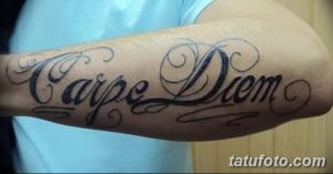 Фото тату надписи 'Carpe Diem' на предплечье, выполненной в черном цвете с декоративными завитушками и узорами. Идеальный эскиз тату для любителей каллиграфии.