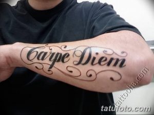 Фото тату надписи "Carpe Diem" с декоративными завитушками на предплечье мужчины. Черно-серый рисунок татуировки, выполненный в каллиграфическом стиле.