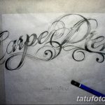 Фотография эскиза татуировки с надписью 'Carpe Diem', выполненного карандашом. Каллиграфический шрифт, детализированная проработка, реалистичный стиль.