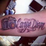 Фото татуировки с надписью 'Carpe Diem' и изображением старинного фотоаппарата. Черно-серый рисунок татуировки на предплечье.