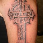 Фото тату креста с надписью 'Carpe Diem' на плече. Черно-серый рисунок татуировки с готическими элементами и латинской фразой.
