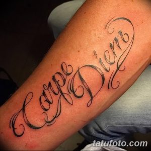Фото тату надписи 'Carpe Diem' на предплечье. Черно-белая татуировка, каллиграфический шрифт, изящные завитки, классический стиль.