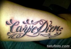 Фото тату: надпись 'Carpe Diem' в классическом стиле на предплечье. Черно-белый рисунок татуировки с декоративными элементами.