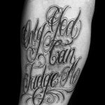 Черно-белое фото татуировки с надписью 'Only God can judge me' на руке мужчины. Изящный шрифт, декоративные элементы, сложный дизайн.