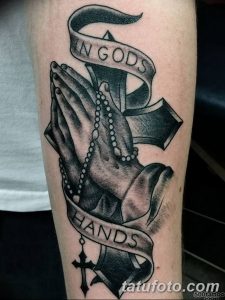 Фото тату на предплечье: молящиеся руки, обернутые лентой с надписью 'In Gods Hands', и крест, выполненное в черно-белой гамме.