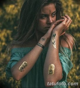 Фото девушки с длинными волосами, украшенной золотыми временными татуировками на руках и запястьях, в окружении зелени.