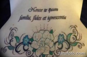 Фото тату с латинской цитатой и цветами на спине. Элегантный рисунок в черно-белой гамме с добавлением голубых акцентов.