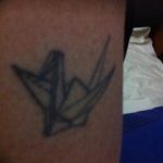 Фото минималистичной татуировки в виде сложенного из бумаги журавлика, выполненной тонкими линиями.