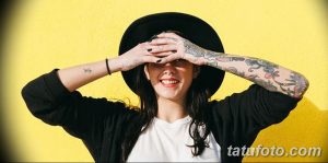 Фото тату девушки с татуировками на руках. Она позирует в шляпе и черной куртке, прикрывая глаза рукой. Детализированное изображение.