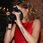 Фото тату: девушка-фотограф с татуировкой в виде надписи на запястье. Она держит в руках фотоаппарат Canon.