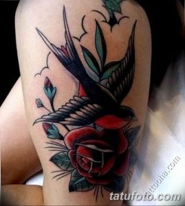 Фото красочной татуировки ласточки, летящей над красной розой на плече. Классический рисунок татуировки в стиле олдскул с яркими цветами и четкими контурами.