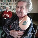 Фото тату: пожилая женщина с небольшой татуировкой в виде спиц и клубка ниток на плече, сидящая в кресле.