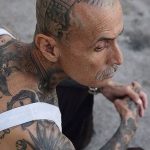 Фото тату: пожилой мужчина с татуировками, покрывающими его голову, шею и плечи, в стиле традиционной тюремной татуировки.