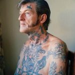 Фото тату: пожилой мужчина с татуировками на шее, руках и груди, изображающими птиц и цветы в синих тонах.