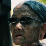 Фото тату: пожилая женщина в очках демонстрирует татуировки на лице, выполненные в темных тонах.