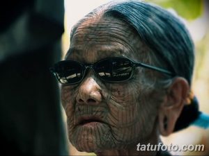 Фото тату: пожилая женщина в очках демонстрирует татуировки на лице, выполненные в темных тонах.