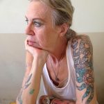 Фото тату: пожилая женщина с татуировками на руке, изображающими черепа, цветы и другие элементы в ярких цветах.
