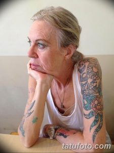 Фото тату: пожилая женщина с татуировками на руке, изображающими черепа, цветы и другие элементы в ярких цветах.