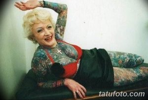 Фото тату: пожилая женщина, покрытая татуировками в стиле олдскул и традиционный, позирует в расслабленной позе.