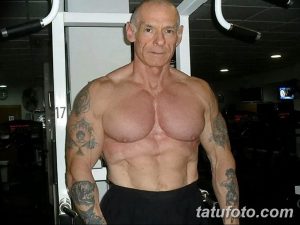 Фотография мускулистого мужчины старшего возраста с татуировками на руках и груди, демонстрирующего свою физическую форму в тренажерном зале.
