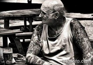Фотография пожилого мужчины с татуировками, сидящего в одиночестве за столом, демонстрирующая его внутренний мир и размышления.
