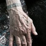 Фото минималистичной татуировки на запястье пожилого человека. Татуировка представляет собой тонкую серую полосу.