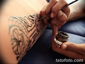 Фото тату: мастер наносит татуировку в стиле трайбл на руку клиента, видны детали процесса и черные узоры.
