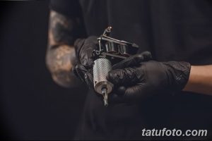 Фото тату: крупный план тату-машинки, удерживаемой в руках мастера в черных перчатках. На руке мастера видны другие татуировки.