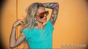 Фотография стильной девушки в голубой майке и с татуировками на руке, позирующей на фоне оранжевой стены.