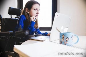 Фотография девушки, работающей за ноутбуком в инвалидной коляске. На столе стоит кружка с рисунком. Иллюстрация современной жизни.