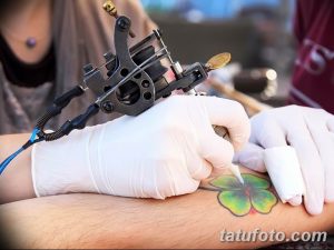 Фото процесса нанесения татуировки клевера на руку, с видимой тату-машинкой и перчатками мастера.