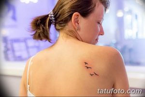 Фото тату с изображением нескольких маленьких черных птиц, летящих вверх, на женской лопатке. Минималистичный рисунок татуировки.