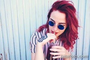 Фотография девушки с татуировкой на руке. Девушка пьет напиток через трубочку и смотрит в камеру.