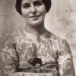 Чёрно-белое фото женщины, покрытой татуировками от шеи до рук. Изображения цветов, бабочек и других узоров.