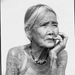 Фотография пожилой женщины с традиционными татуировками на лице и теле. Рисунок выполнен в этническом стиле.