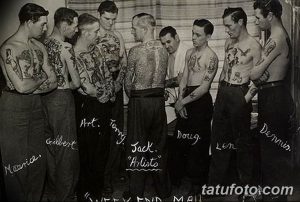 Чёрно-белая групповая фотография мужчин с татуировками, сделанными в старом тату-салоне. Видны тату-мастера и клиенты.