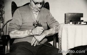 Фотография пожилого тату-мастера, работающего над татуировкой. Видно тату-оборудование и татуировки на руках мастера.