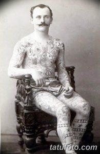 Старинная фотография мужчины, полностью покрытого татуировками. Мужчина сидит в кресле и демонстрирует свои татуировки на руках, груди и ногах.