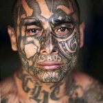 Фотография мужчины с полностью покрытым татуировками лицом и телом. Татуировки выполнены в стиле традиционной тюремной татуировки, с использованием черных и синих чернил.