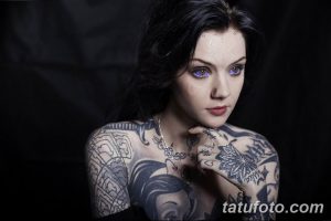 Фотография девушки с темными волосами и голубыми глазами, покрытой сложными татуировками на плечах, груди и руках. Тату выполнены в темных тонах.
