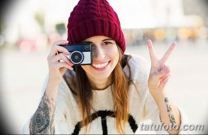 Фотография молодой девушки с татуировками на руках, держащей старинную камеру и показывающей жест 'V'