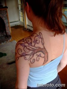 Фото тату: красивое дерево с цветущими ветвями на плече девушки. Рисунок выполнен в мягких коричневых и фиолетовых тонах.