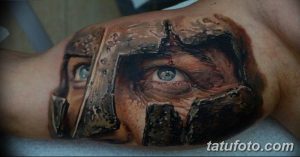Фото реалистичной татуировки на плече, изображающей шлем древнегреческого спартанца. Детализированная проработка металла и глаз.