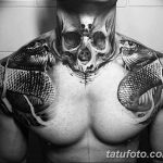 Фотография татуировки на груди и плечах с изображением черепа, обвитого двумя змеями. Детализированный рисунок в стиле реализм.