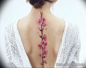 Фото акварельной татуировки с изображением ветки цветущей сакуры на спине девушки. Нежные розовые оттенки и размытые контуры создают эффект акварельной живописи.