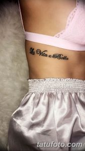 Фото тату надписи «La Vita e Bella» на боку женщины. Черная, изящная каллиграфия, выполненная вдоль линии талии. Изящный рисунок татуировки.