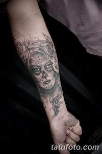 Фото тату сахарной черепушки (Santa Muerte) с цветами и детальной прорисовкой на предплечье. Рисунок выполнен в черно-белых тонах с яркими акцентами.