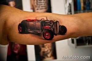 Фото тату реалистичного изображения старинного автомобиля на предплечье. Детализированный рисунок с использованием черных и серых оттенков, с красными акцентами на колесах.