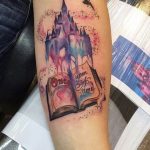 Фото тату на руке: красочный рисунок замка из сказки, открытой книги и летящей птицы, выполненный в акварельном стиле.