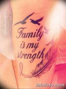 Фото татуировки на плече с надписью "Family is my strength", изображением двух птиц и пера. Черно-белый рисунок татуировки выполнен в реалистичном стиле.
